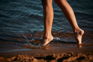 A barefoot girl enters the water to swim. Legs of a slender girl close-up. Rest at a resort near the sea, river. Beach season. Splashes are flying from under your feet