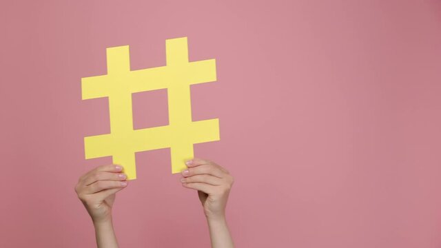 Close up of woman hands hold large yellow paper hashtag symbol, shaking left and right, hash sign of famous media content, social media marketing and blog promotion, isolated on pink studio background