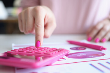 Woman with pink manicure counting on calculator near paper with charts closeup