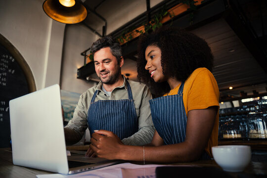 Manager Of Coffee Shop Training Intern Showing Menu On Laptop While Drinking Hot Coffee In Cafe 