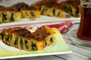 A slice of spinach patty on a dessert plate. A glass tea cup with tea on the right and blured view of slices of spinach paty on serving plate behind.