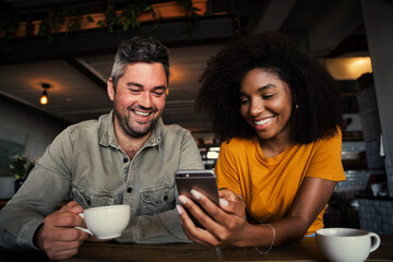 Mixed race couple giggling at pictures on social media holding smartphone in trendy cafe shop