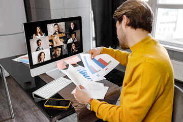 Concentrated male employee in yellow shirt working from home, having online briefing meeting with colleagues. Serious young businessman doing paperwork, learning financial graphs, distant work concept