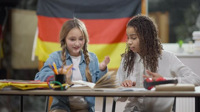 Smart genius Caucasian schoolgirl writing in workbook and declaiming as African American classmate entering classroom with German flag at background. Intelligent girls in junior high school