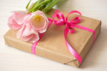 Gift wrapped in craft paper with a pink ribbon and a bouquet of delicate pink tulips. Small cute gift close-up