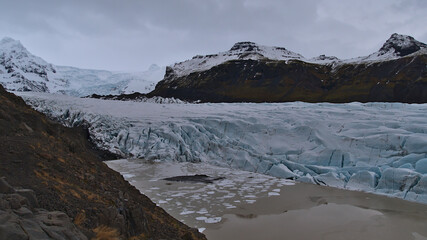 View of the breakoff edge of mighty Svínafellsjökull glacier, part of Vatnajökull ice cap in southern Iceland, with crevasses, glacial lake and rugged mountains on cloudy winter day.