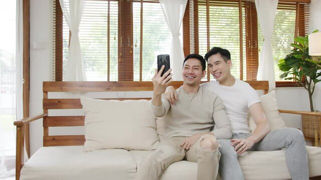 Happy young asian gay couple sit couch use smartphone facetime video call with friends and family in living room at home. Stay at home quarantine, Social distancing, LGBTQ+ Young married concept.