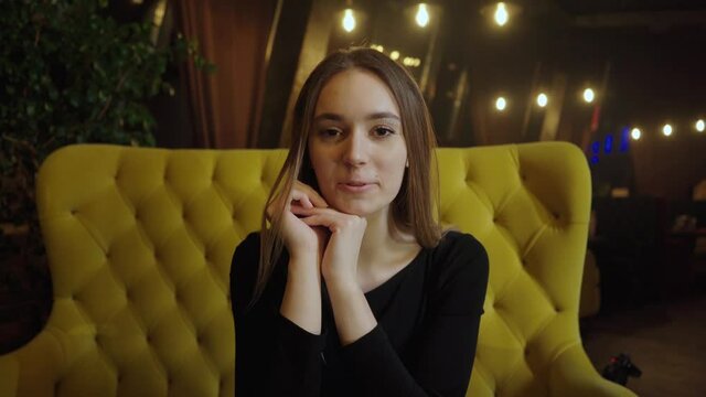 A young girl in a black dress sits in a coffee shop in the evening on a yellow sofa and talks, looking directly into the camera