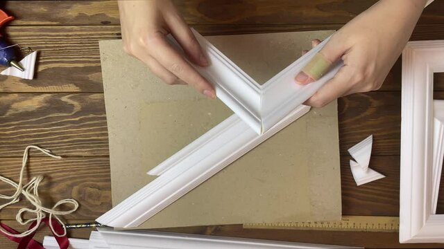 Women's hands gluing details of frames made of styrofoam. Interior decorations made of moldings. Diy handicraft process. Wooden table, hot glue and cardboard on background. Designer's workspace.