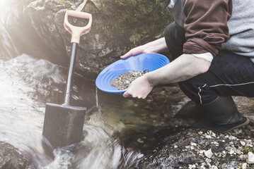 Outdoor adventures on river. Gold panning, search for gold. Man is looking for gold with a gold pan in a small stream. Dark moody look - obrazy, fototapety, plakaty