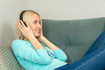 Young blonde woman in bright clothes is lying on the sofa with large headphones listening to music and smiling