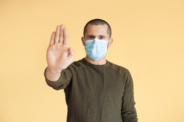 Short haired man is gesturing the stop sign with palm posing on a yellow studio wall wearing a medical mask