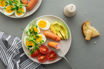 Delicious breakfast, boiled eggs with sausages, tomatoes and avocado, microgreens of peas and sunflowers, plates with food on a gray background ,top view