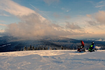 Two people on snowmobiles in a snowy clearing.