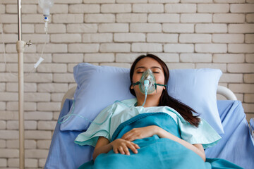 Young long black hair sick asian female patient wear green shirt and oxygen intubation mask on face sleeping on blue sheet hospital pillow and bed in icu ward room with white brick wall background - obrazy, fototapety, plakaty