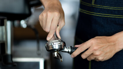 close up photo of male hands holding a metal tamper and a portafilter with coffee in a coffee shop. A man barista preparing for pressing ground coffee for brewing espresso or americano in a cafe.