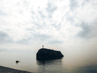 A huge rock in the sea with a cross, against the background of a cloudy sky merging with the...