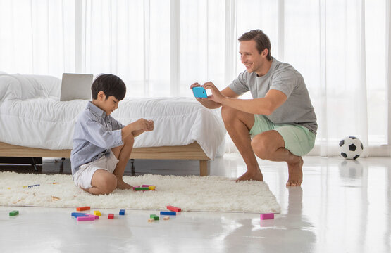 Father using smartphone to take photos of his mixed-race son who playing with a colorful wooden block in home bedroom. Idea for lifestyle of working from home