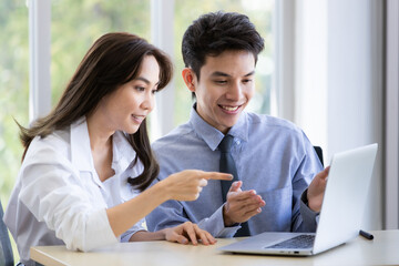 Beautiful black long hair Asian businesswoman and gook looking cute Asian businessman sitting together and watching the screen of laptop notebook computer in office with smile face