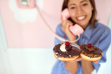 Closeup of a young beautiful girl who is having an interesting conversation on the phone and posing for a photo in a retro looking pastry shop while holding chocolate donuts in her hand