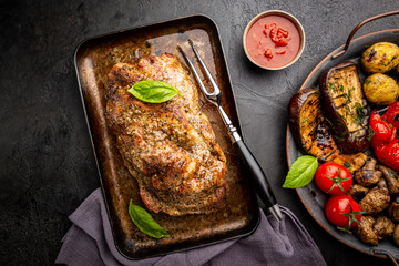 Baked pork meat with grilled vegetables in a baking pan on dark background, top view