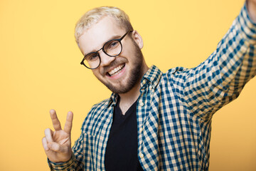 Naklejka premium Caucasian man with beard and blonde hair is making a selfie on a yellow studio wall smiling and gesturing peace sign