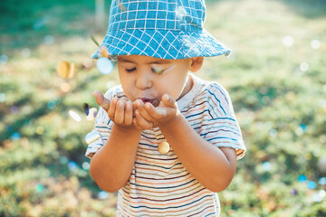 Small caucasian boy is blowing confetti from his palm playing in the garden wearing a blue hat