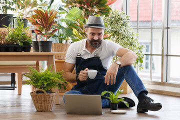 A bearded man wearing hat sitting and drinking coffee and using laptop notebook computer to sell plant in home indoor garden. Online marketing and live stream seller concept