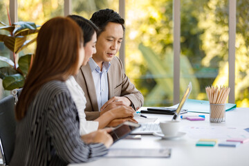 young attractive Asian businesswoman dressed formally sitting and holding tablet with smiley face during working in the office with colleagues sitting in the background