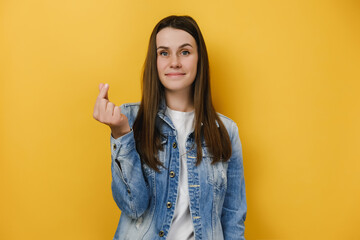 Portrait of dreamy beautiful young European woman makes love sign, shapes mini heart with fingers, feels happy, wears blue denim jacket, isolated on yellow studio background. Body language concept