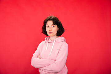 Young beautiful brunette woman in trendy pink hoodie looks at camera with crossed arms isolated on...