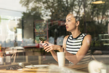 Thoughtful caucasian woman holding mobile phone while looking through the coffee shop window during coffee break.