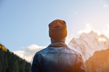 Fototapeta na wymiar View from the back. A man in a denim jacket and a brown hat stands in the forest in the mountains against the background of the evening sky and snow-capped mountain peaks