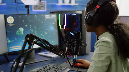 Pro cyber black woman streaming videogame, reading on stream chat and playing space shooter on RGB powerful computer. Streamer sitting on gaming chair playing game during online esports tournament. © DC Studio