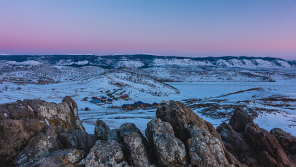 Fototapeta na wymiar Dawn over a snowy valley. The sky above the ridge turns pink. Village houses on the shore of a frozen lake. In the foreground there are picturesque rocks. Stone texture. Baikal