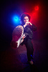 A brunette curly girl in black shiny clothes looking as teen bully on a black background. Model and actress posing in the studio