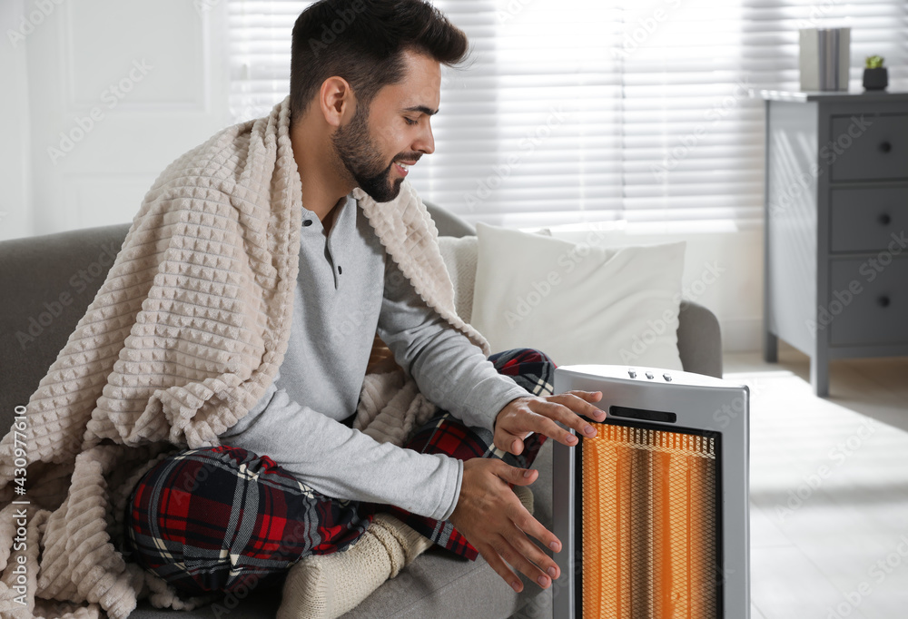 Sticker Young man warming hands near electric heater at home