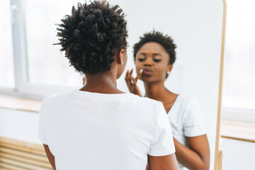 Portrait of happy beautiful young African American woman in white t-shirt takes make up near mirror