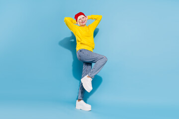 Full size photo of cheerful joyful old woman hold hands behind head wear hat isolated on blue color background