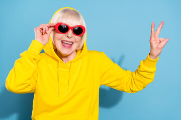 Photo of cheerful nice happy old woman wear sunglass hood make v-sign isolated on blue color background