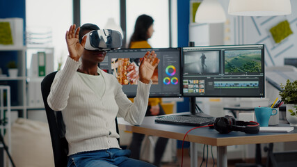 African video editor experiencing VR goggles, gesturing, editing video film montage working with footage and sound on computer with two displays. Videographer processing movie in creative agency