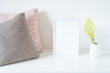 Empty white frame mockup on white table. Soft colored cushions and white vase with an arum italicum...