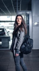stylish young girl with long dark hair in a gray sweatshirt with a hood and a black leather backpack in a hangar or large garage. female student in a gray coat in an industrial area