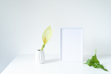 Empty white frame mockup on white table. Modern white vase with arum italicum flower and green...