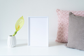 Empty white frame mockup on white table. Soft colored cushions and white vase with an arum italicum...