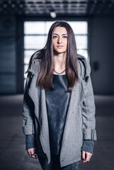 Young girl with makeup and long dark hair in a gray coat with a hood and a black leather backpack in a hangar or large garage