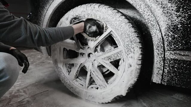 Hands of young man holding special cleaning glove, washing car wheel with foam. Cleaning of modern rims of luxury car at self car wash service.