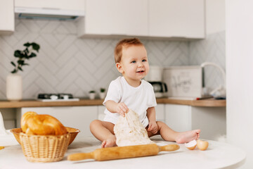A little boy is sitting on the kitchen table with a rolling pin and a baking dough. Mom's assistant, baby, childhood