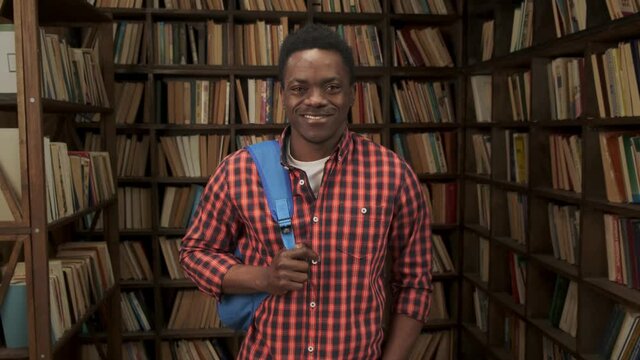 African American male student holding backpack in university or high school library smiling looking at camera portrait
