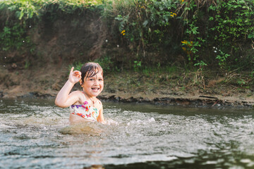 Fototapeta premium Happy summertime, healthy childhood concept. Little girl swimming, splashing, jumping and having fun in a river in summer. Blured green background. Horizontal shot.